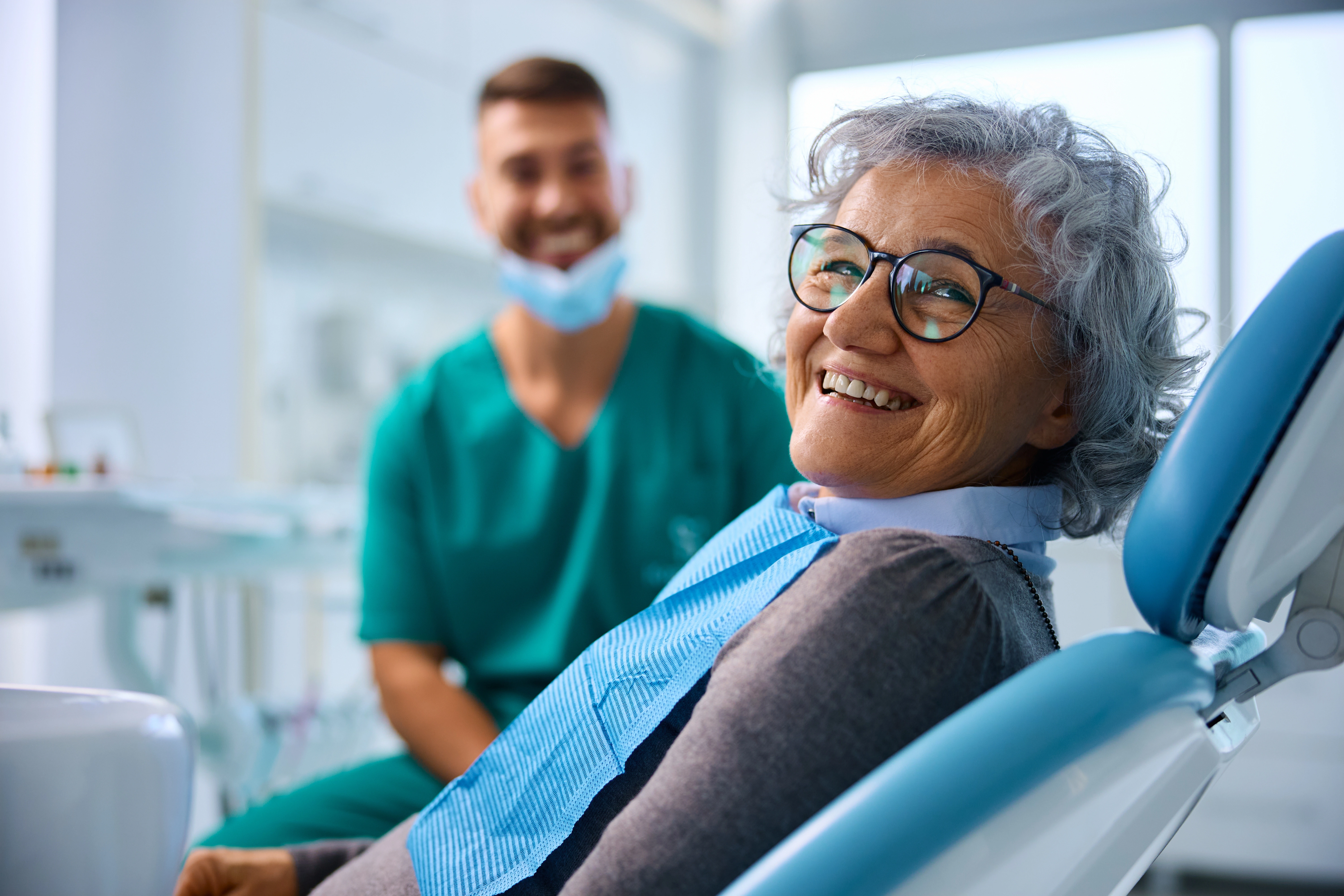 A cheerful senior woman smiles while seated in a dental chair, with her dentist in scrubs smiling in the background.
