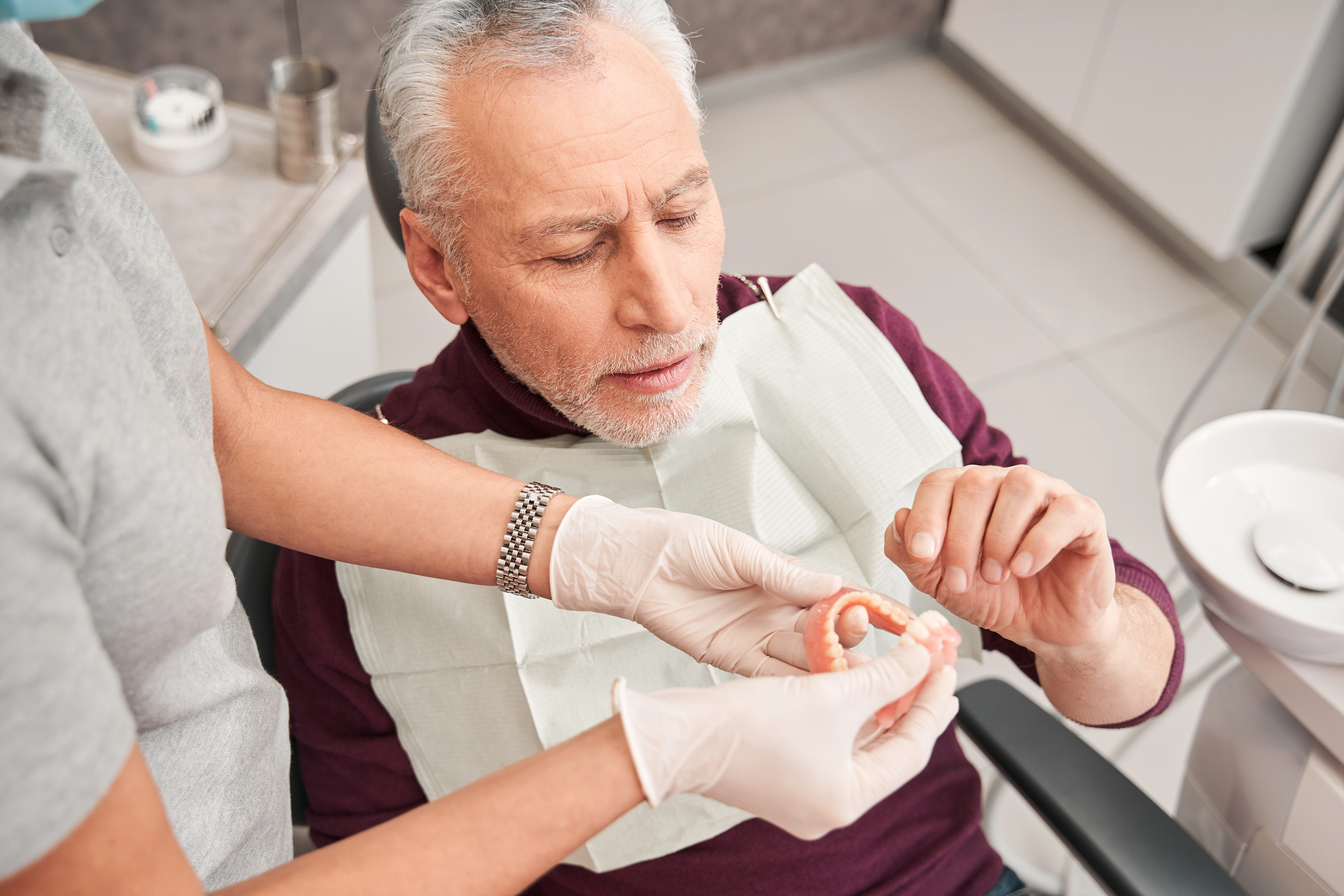 An elderly man in a dental chair examines a set of lower dentures handed to him by a dentist wearing gloves, likely discussing fit and functionality.