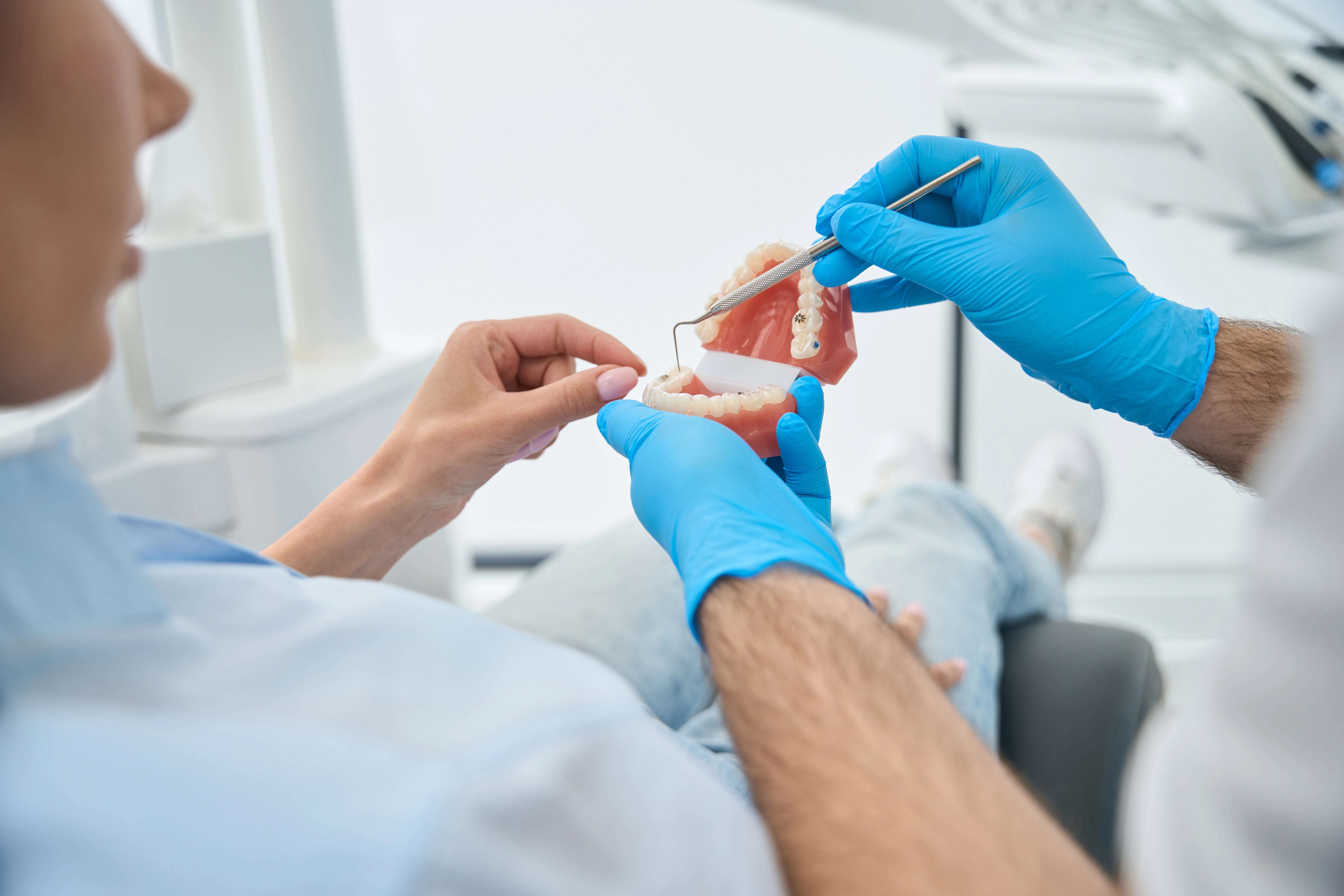 A dentist wearing blue gloves uses a dental tool to demonstrate on a model of teeth, pointing to the lower dentures while a patient observes closely during a consultation