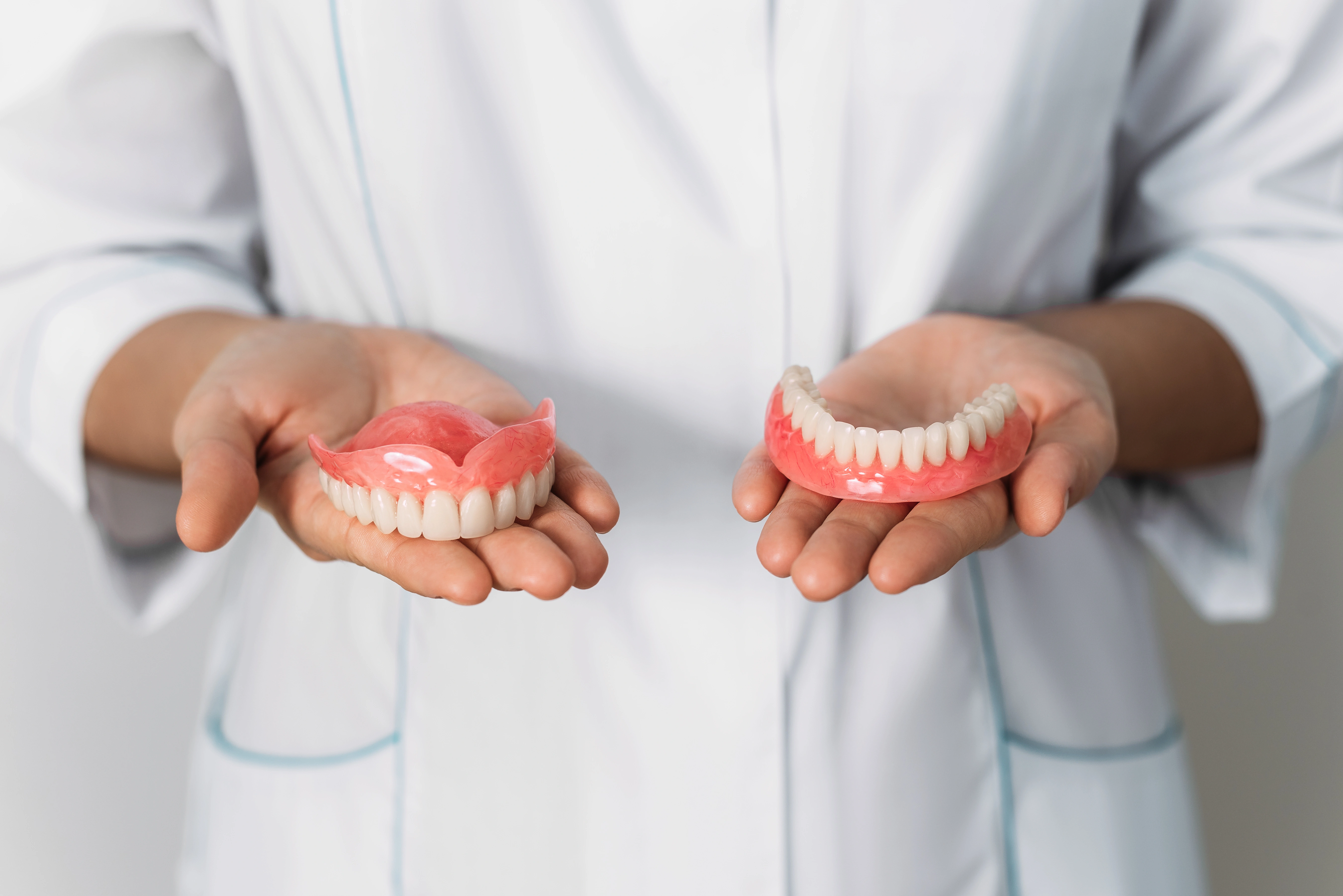 A dentist in a white coat holds a pair of upper and lower dentures in their hands, likely demonstrating the difference between the two or discussing denture care.