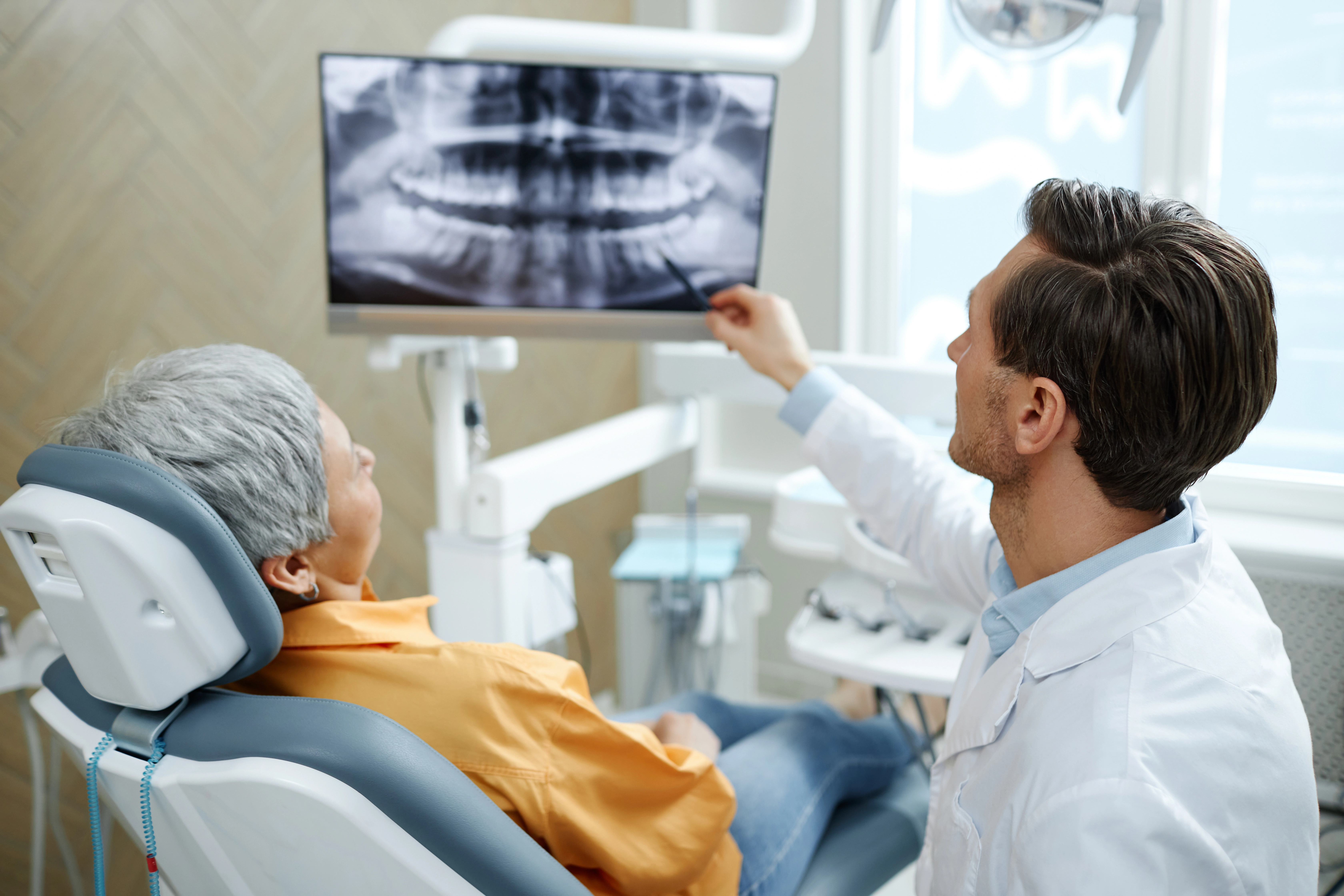 A dentist reviews a dental X-ray displayed on a monitor with a patient seated in the examination chair, explaining the findings.